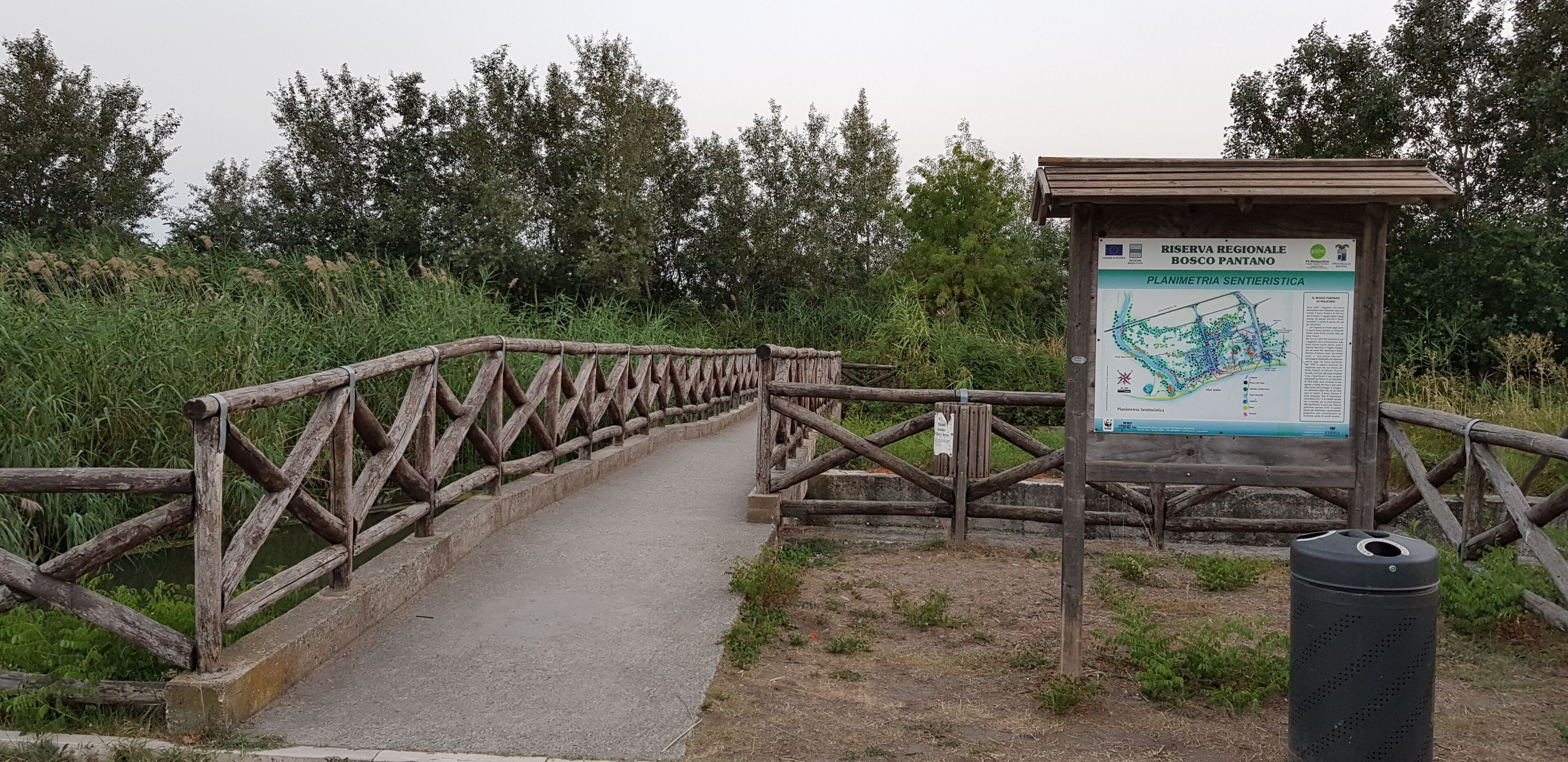 Un pont en bois, agrémenté d’un panneau indicateur, entouré de verdure.