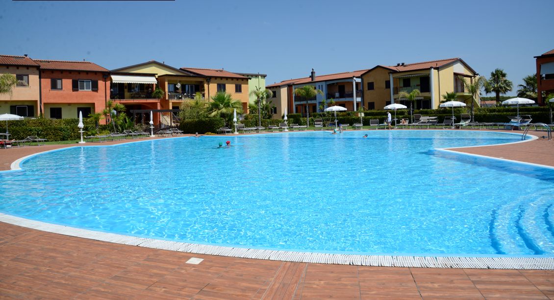 Uma grande piscina com um deck revestido, rodeada por edifícios com varandas e guarda-sóis.