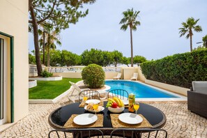 A poolside dining area with a table set for two, including plates, cutlery, and a fruit bowl. - Villa Vista Mar (Loulé)