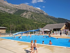A swimming pool area with a water slide, surrounded by mountains and a building with a sloped roof.