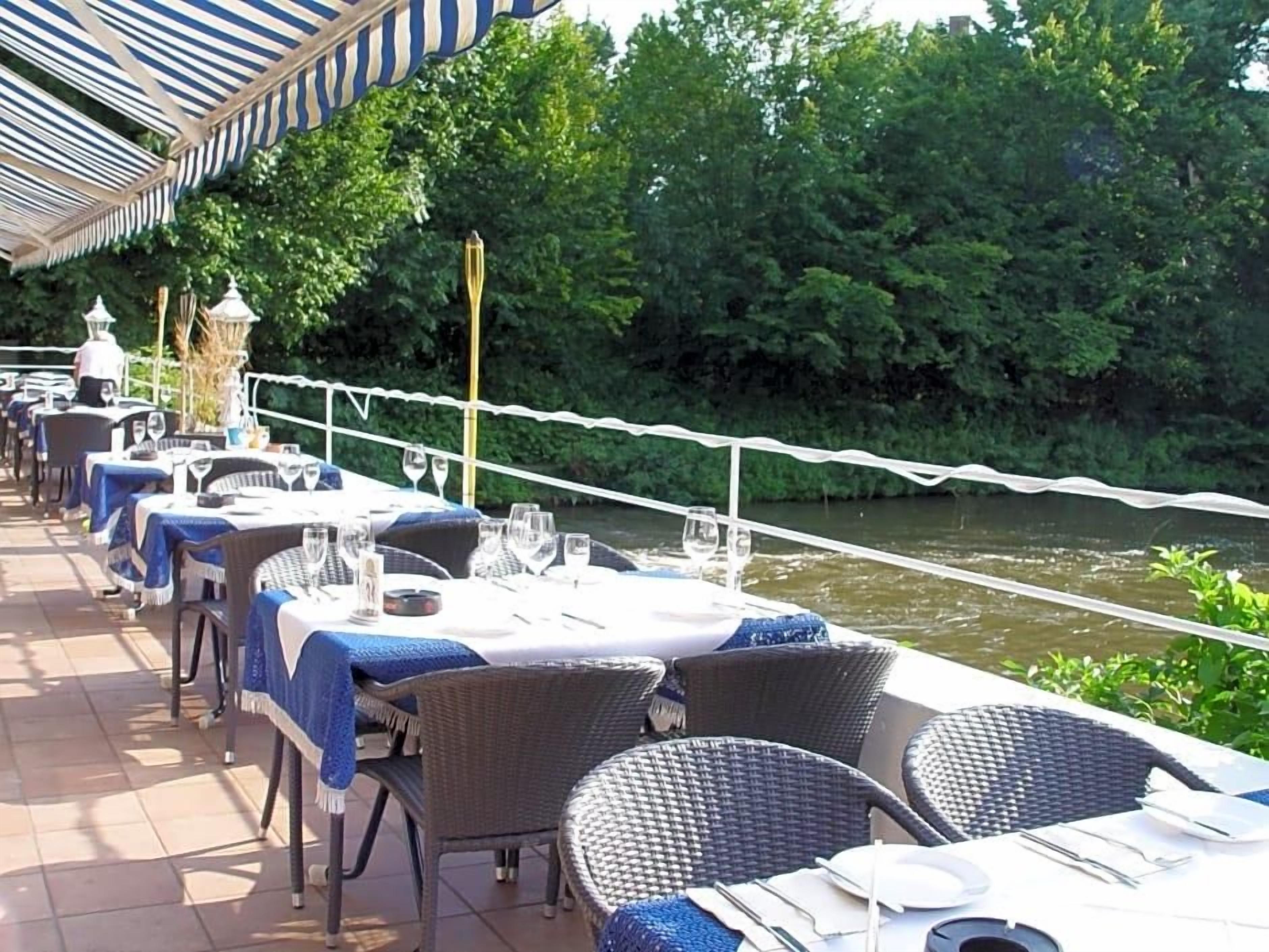An outdoor dining area with tables and chairs, a river view, and a white rope barrier.