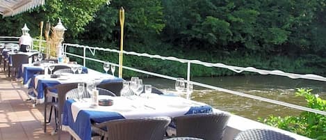 Un área de comedor al aire libre con mesas y sillas, vista al río y una barrera de cuerda blanca.