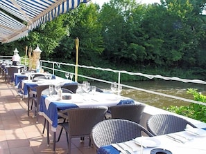 An outdoor dining area with tables and chairs, a river view, and a white rope barrier.