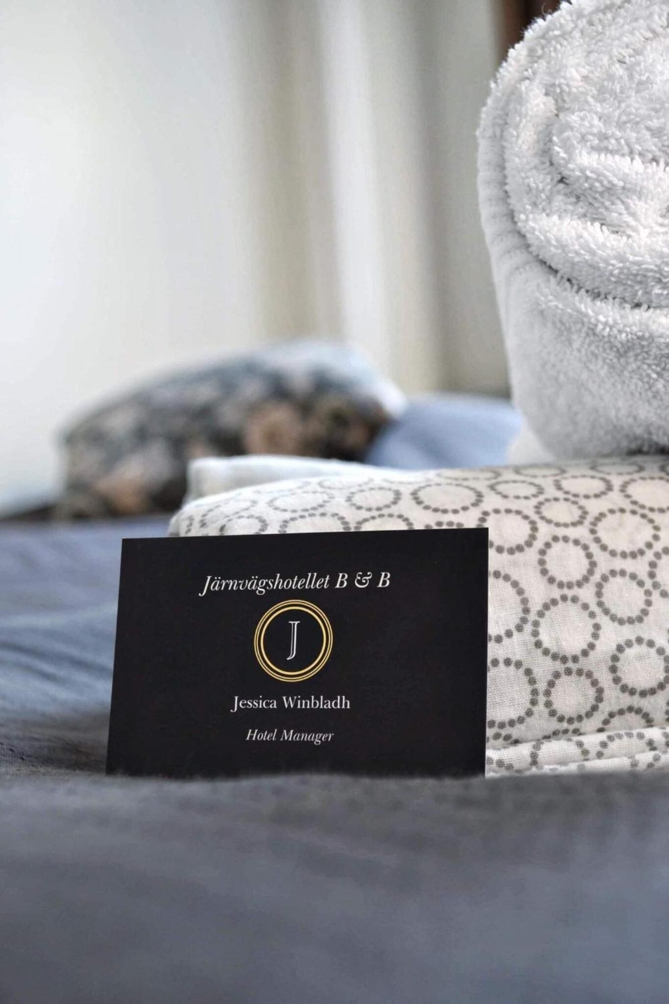 A black business card with the text "Järnväghotellet B&B" and "Jessica Winbladh, Hotel Manager" on a bed with patterned bedding and a rolled white towel.