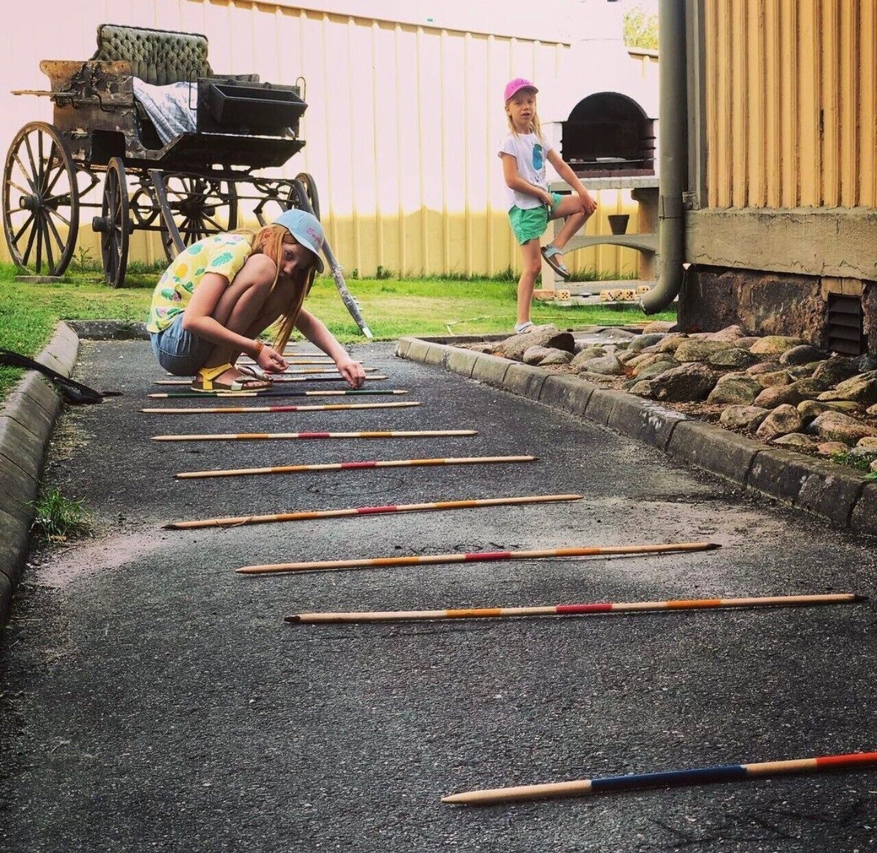 A paved outdoor area with wooden sticks laid out in rows, a vintage carriage, and a child standing by a building.