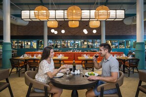 A couple dining at a round table with wicker chairs, surrounded by other tables and chairs, in a restaurant with a brick wall and hanging wicker lamps. - El Beso Adults Only At Ocean El Faro - All Inclusive (Punta Cana)