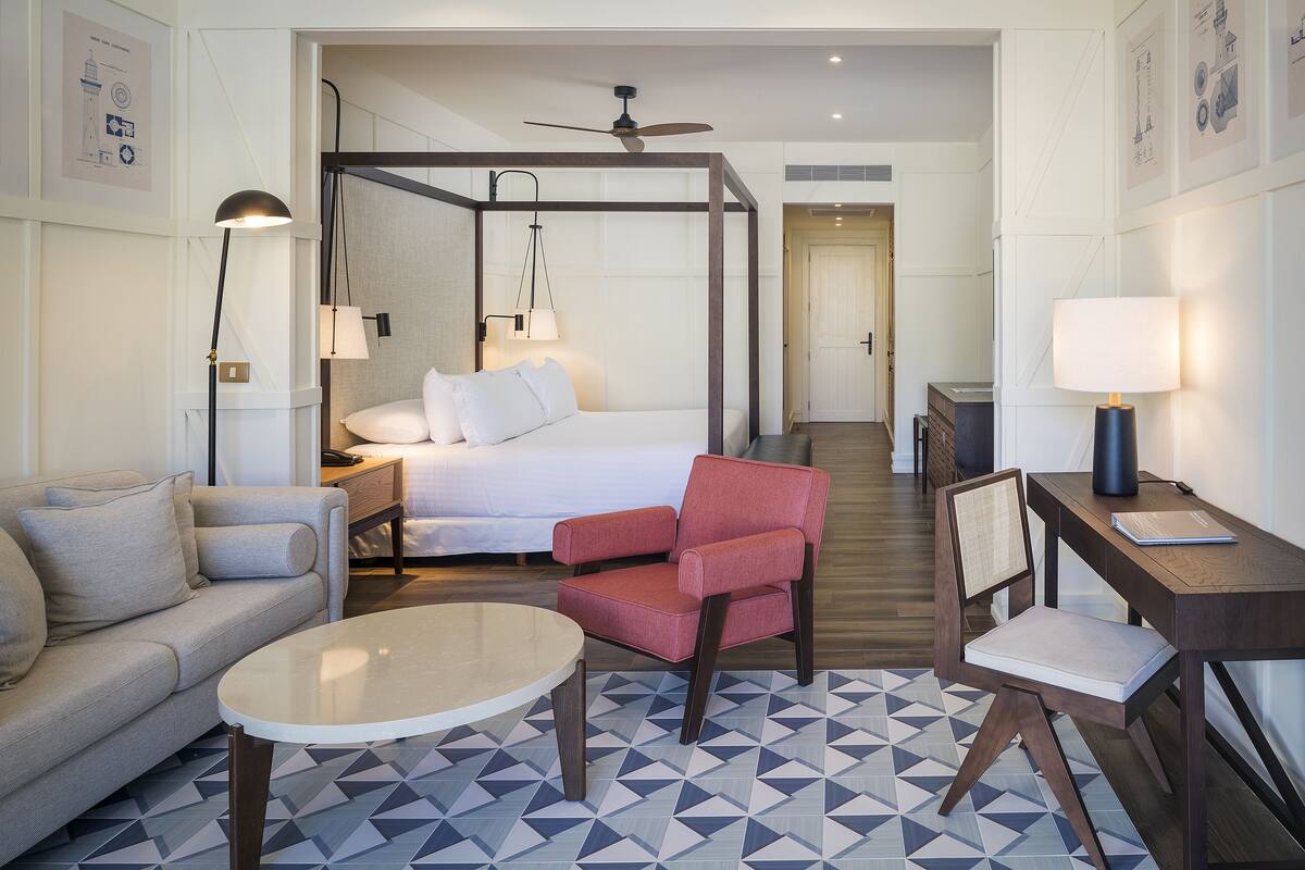A hotel room with a bed, a red armchair, a sofa, a desk, and a coffee table.