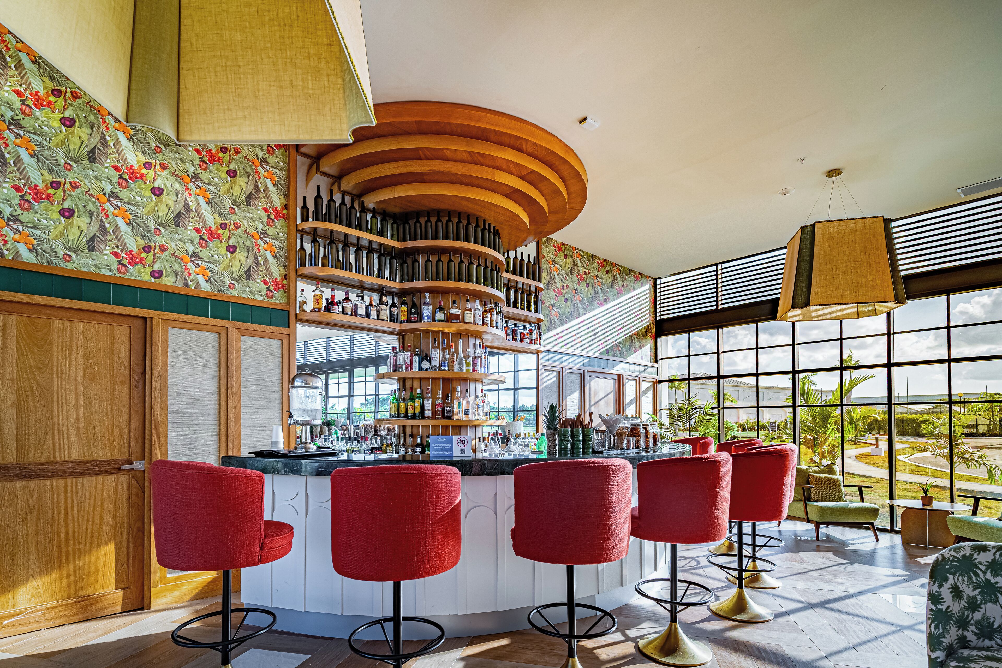 A bar area with a curved wooden shelf, red bar stools, and a view of the outdoors through large windows.