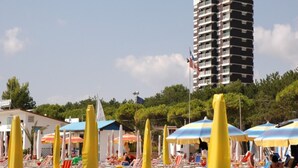 A beach with rows of sun loungers and umbrellas, a tall building in the background, and a clear sky. - Seafront flat with amazing view (Lignano Sabbiadoro)