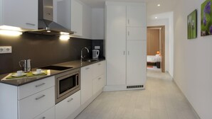 A modern kitchen with white cabinets, a stainless steel range hood, a built-in oven, and a sink. There is a hallway leading to a bedroom with a bed and a nightstand.