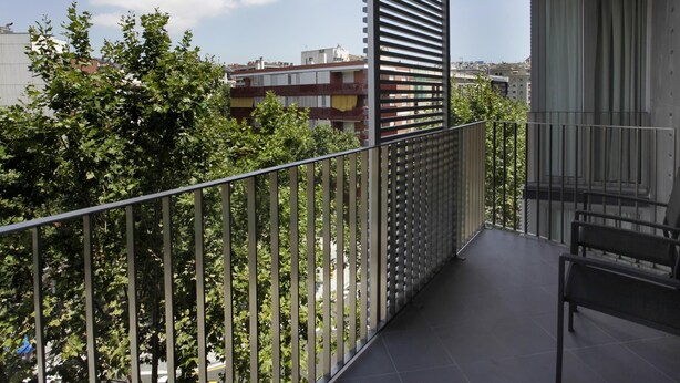 Un balcón con barandilla de metal, una banca y vistas a árboles y edificios al fondo.