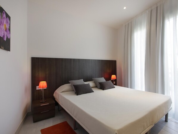 Ein modernes Schlafzimmer mit einem großen Bett, zwei Nachttischen mit Lampen und Blick auf eine Stadtkulisse durch transparente Vorhänge.