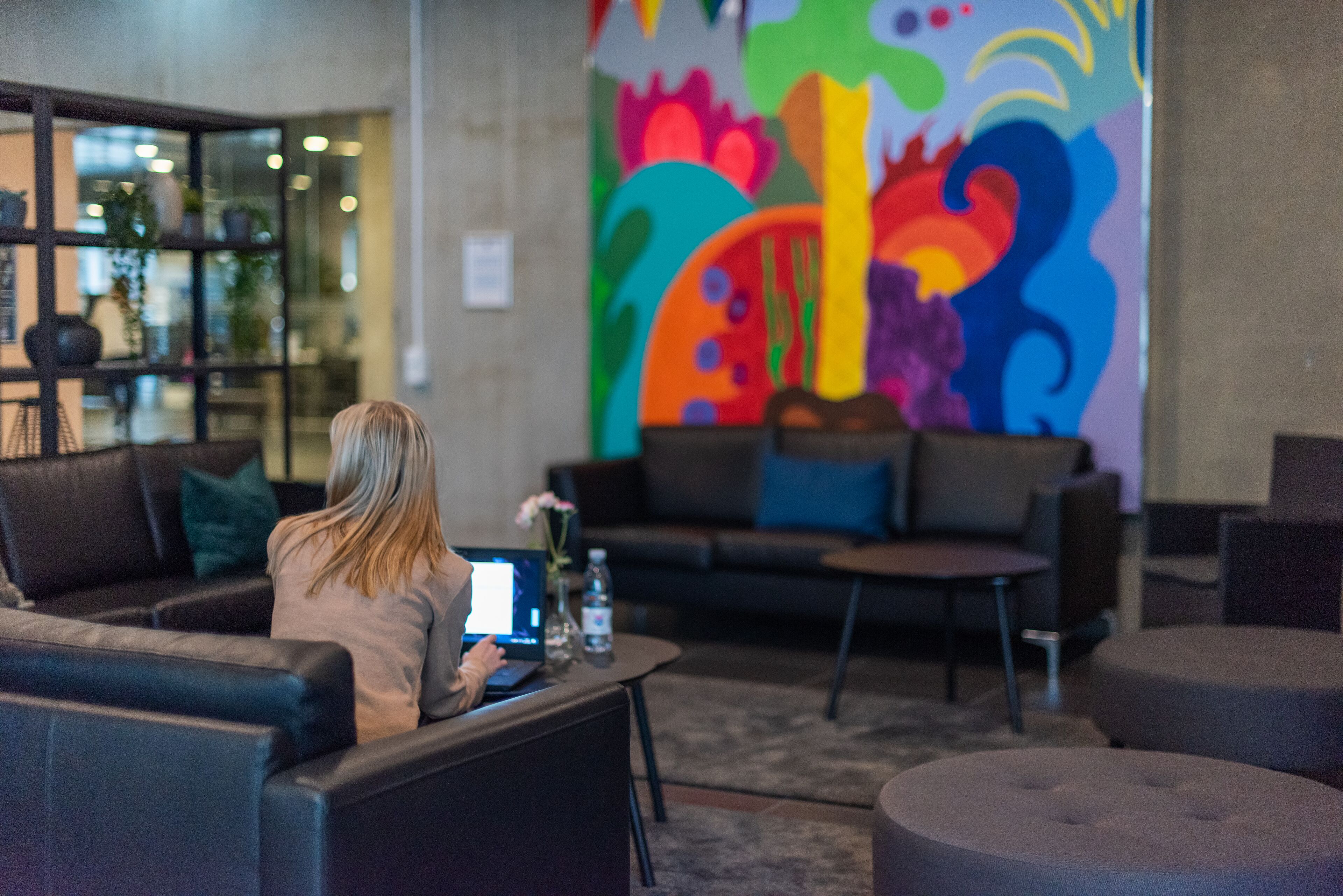 A person using a laptop in a modern lounge area with a colorful mural, comfortable seating, and a glass partition with plants.