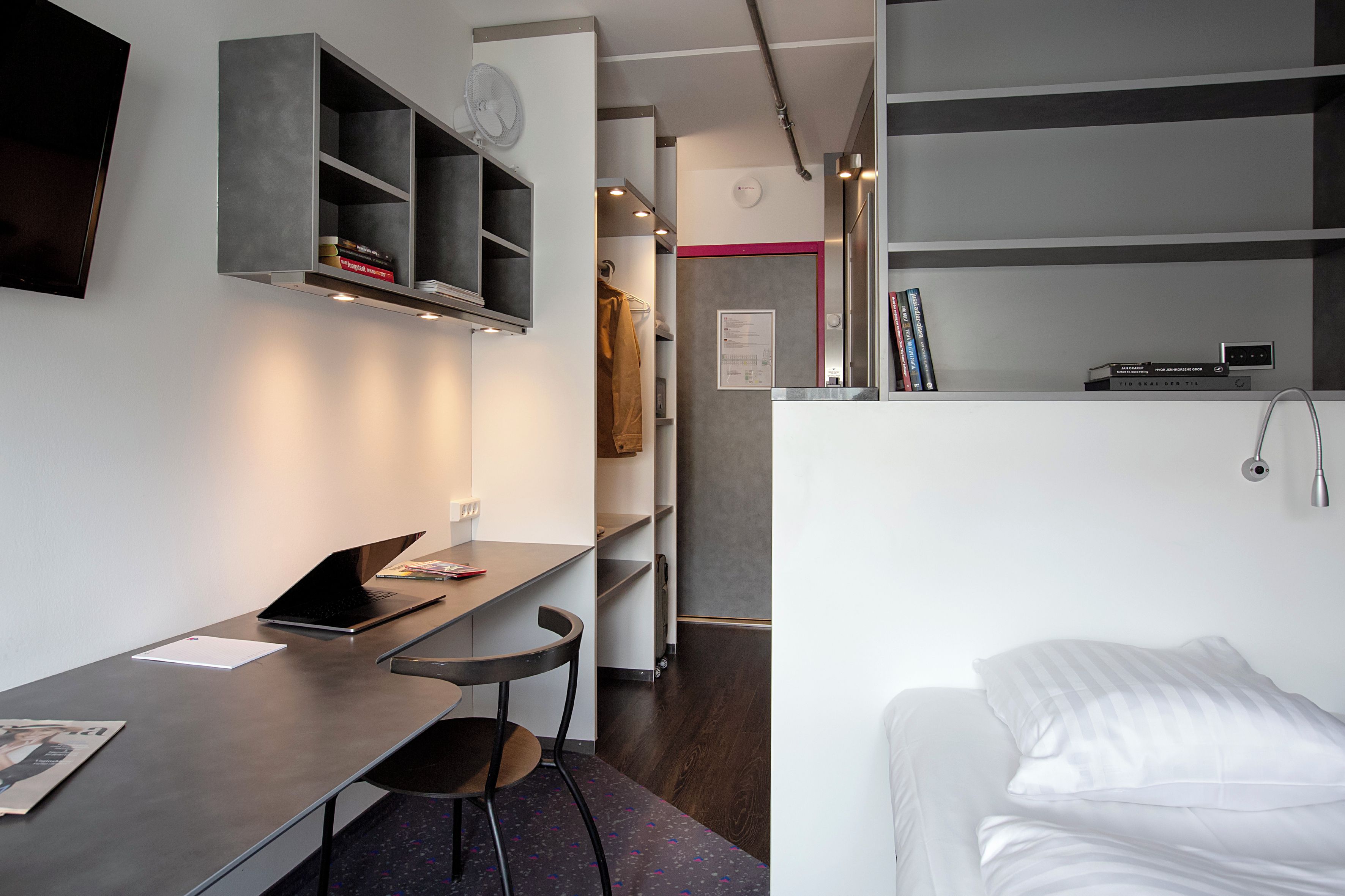 Habitación compacta con cama, escritorio con una computadora portátil, una silla, un estante pequeño con libros y un ventilador de pared.