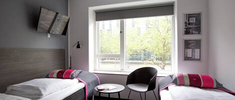 A hotel room with two beds, a small table, a chair, a TV mounted on the wall, and a window with a view of trees and buildings.