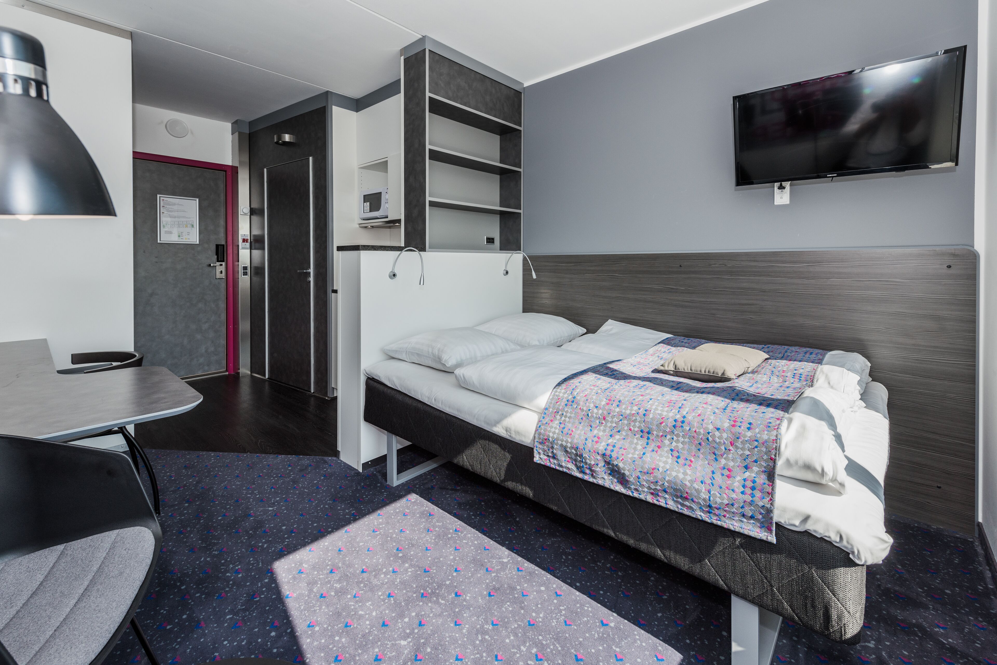 A modern hotel room with a bed, a desk, a chair, a TV, and a door leading to a bathroom.