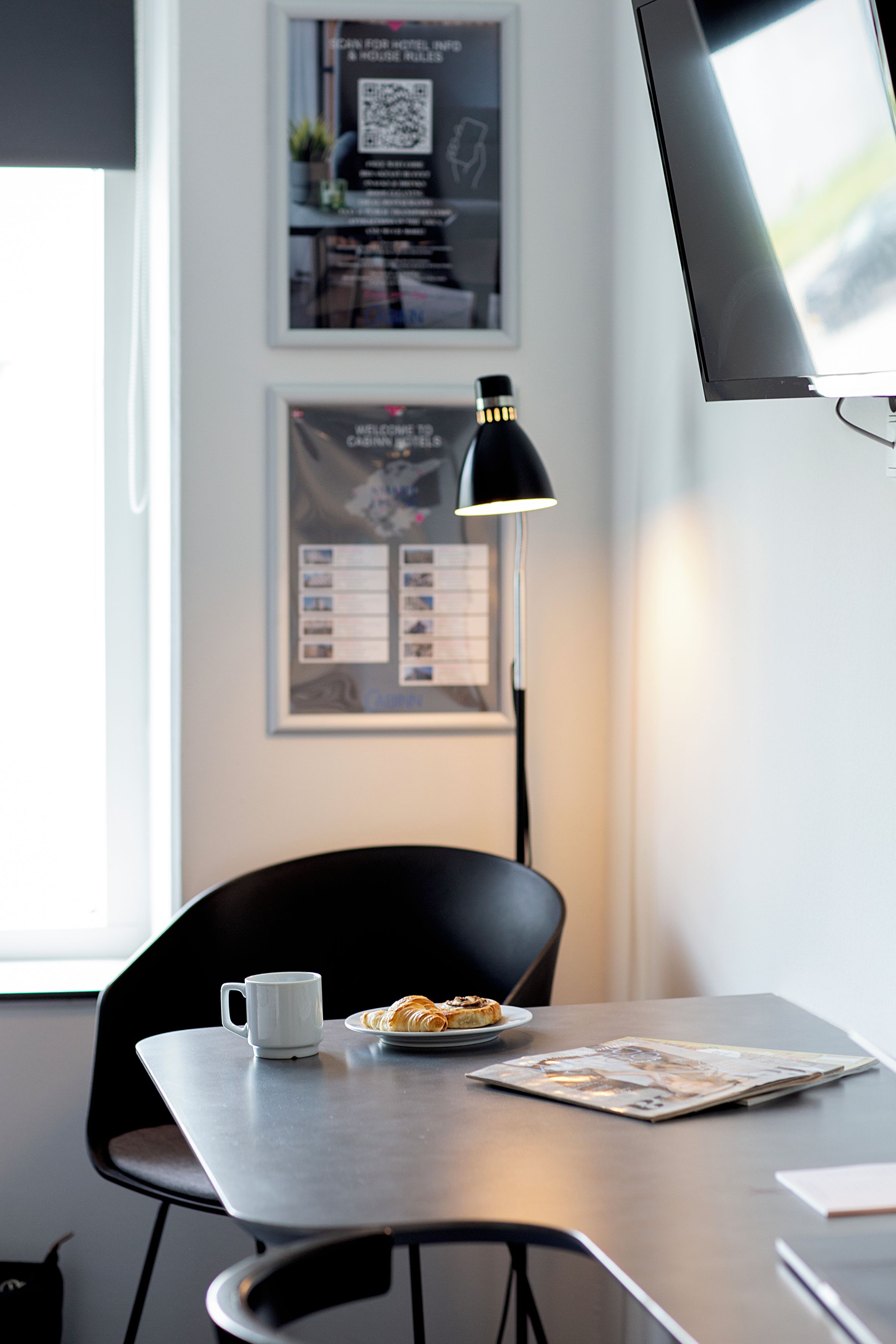 Een moderne kantoorruimte met een bureau, een stoel, een koffiekop, een bord croissants, een lamp en ingelijste informatieve posters aan de muur.