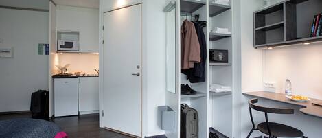 A compact hotel room with a kitchenette, a wardrobe with hanging space, a small table with a chair, and a bed.