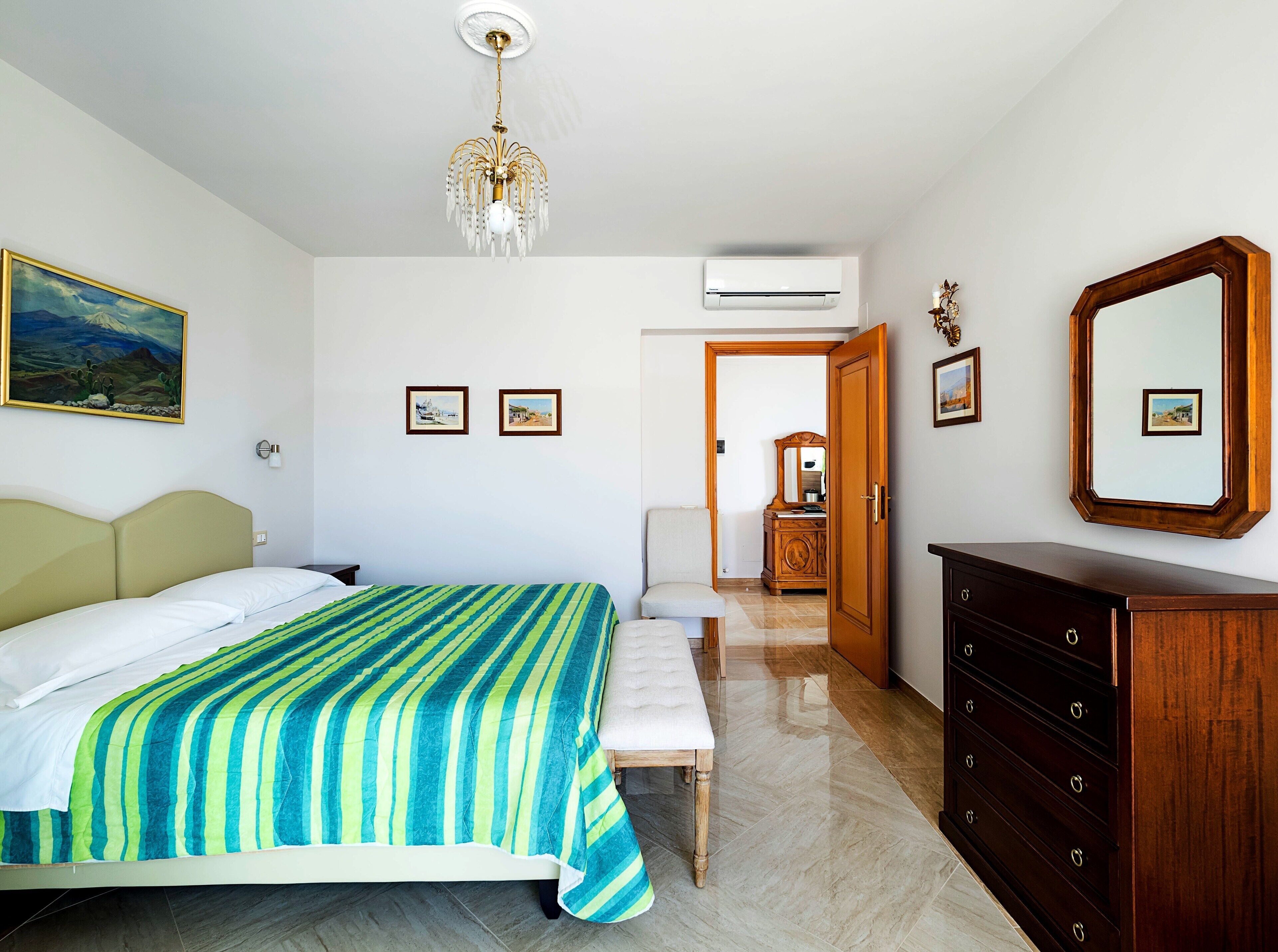 A hotel room with a bed, a bench, a dresser, and a mirror. There are framed pictures on the wall and an air conditioning unit above the door.