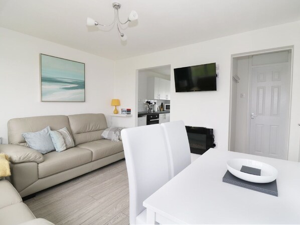 A modern living room with a beige sofa, a white dining table, and a TV mounted on the wall.