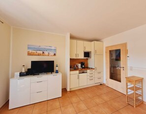 A compact kitchen with white cabinets, a microwave, and a coffee maker. There's a television on a cabinet, a painting on the wall, and a wooden shelf by the door.