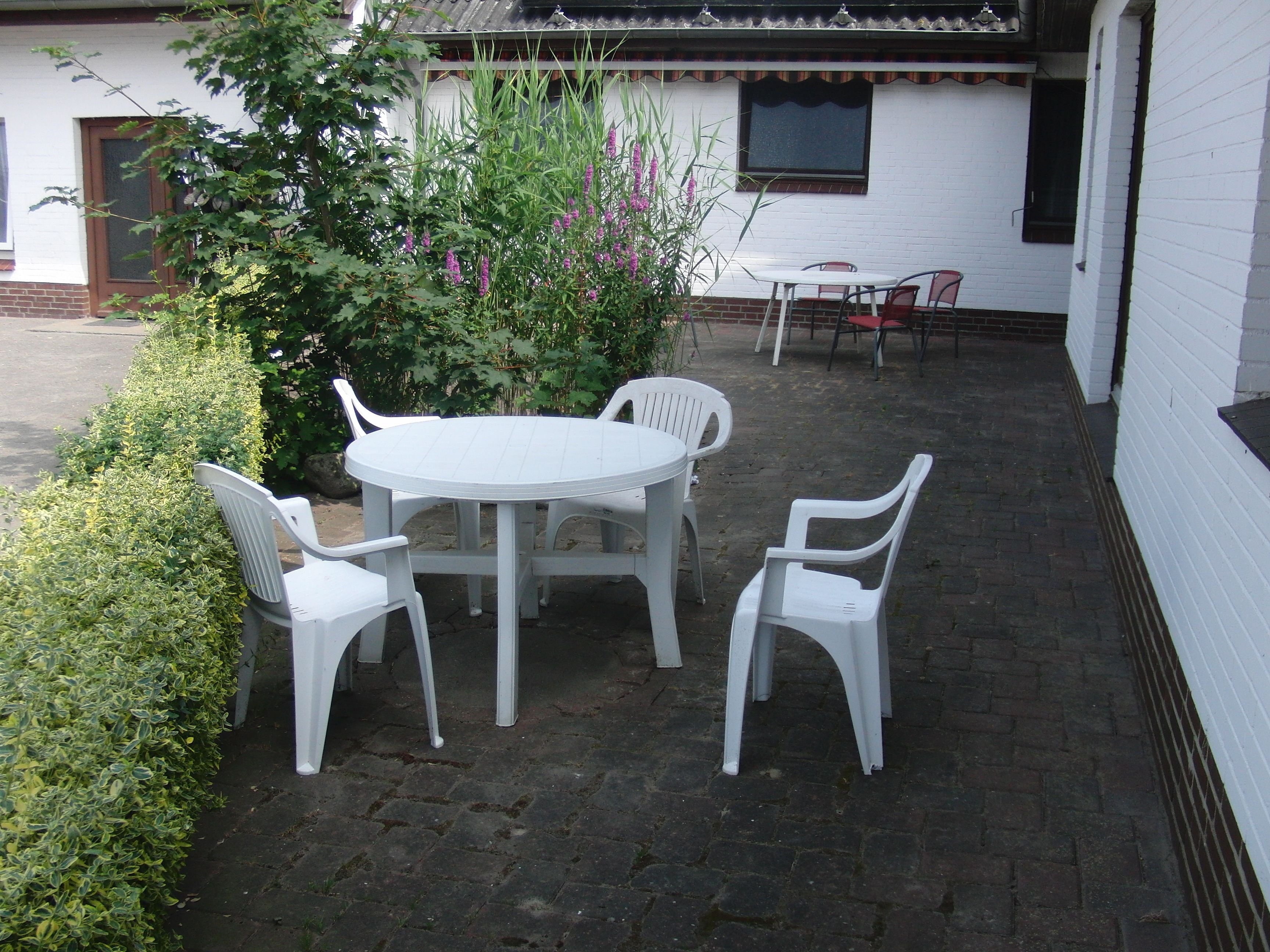 A patio with a round table and four white chairs, surrounded by greenery and a white building with a window and a door.