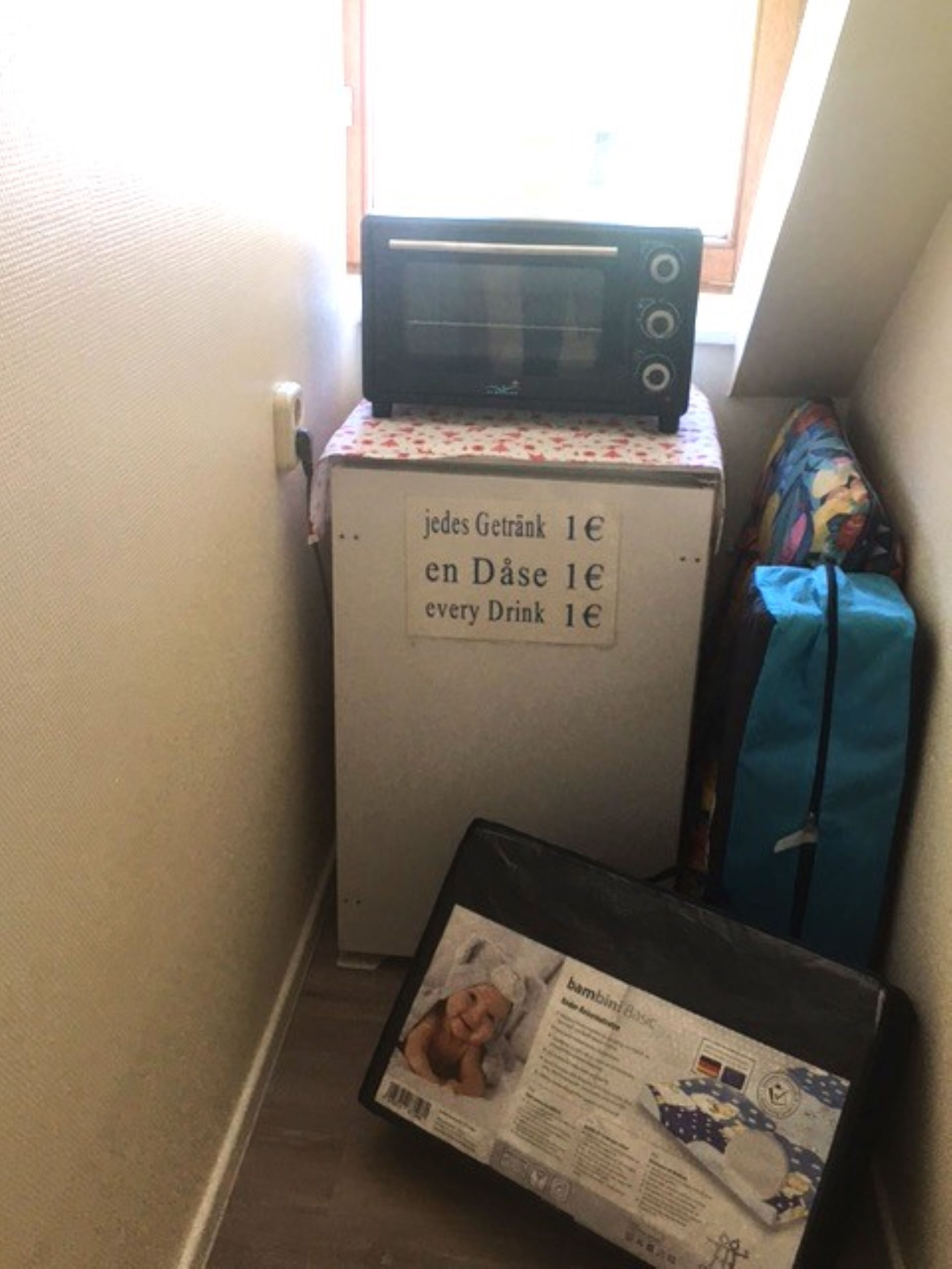 Une petite cuisine équipée d’un micro-ondes posé sur un réfrigérateur, une table recouverte d’une nappe à motifs floraux et un sac bleu sur le sol.