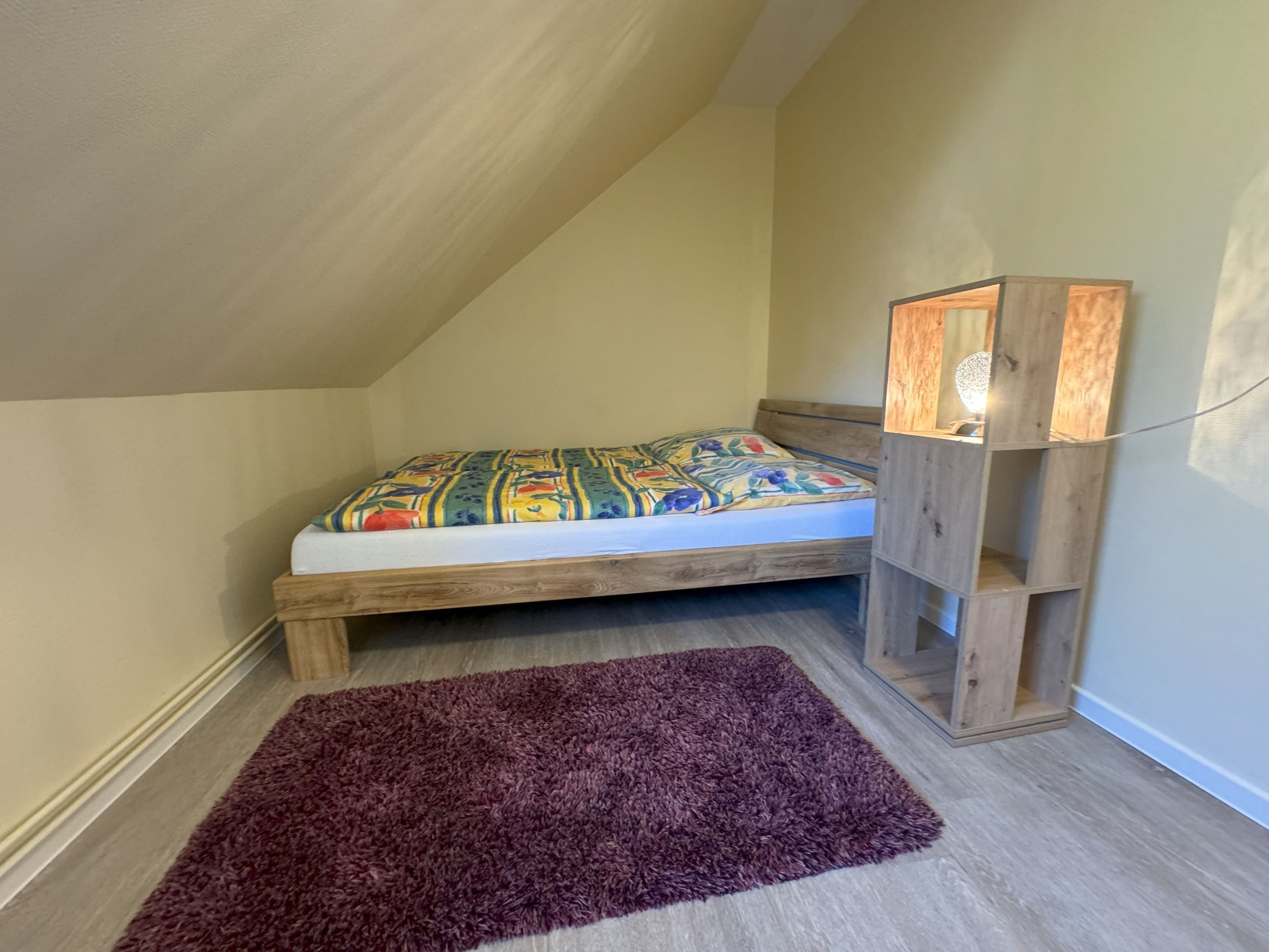 Una habitación pequeña y acogedora con techo inclinado, estructura de cama de madera, colcha colorida, alfombra morada y mesita de noche de madera con lámpara.