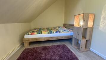 Una habitación pequeña y acogedora con techo inclinado, estructura de cama de madera, colcha colorida, alfombra morada y mesita de noche de madera con lámpara.