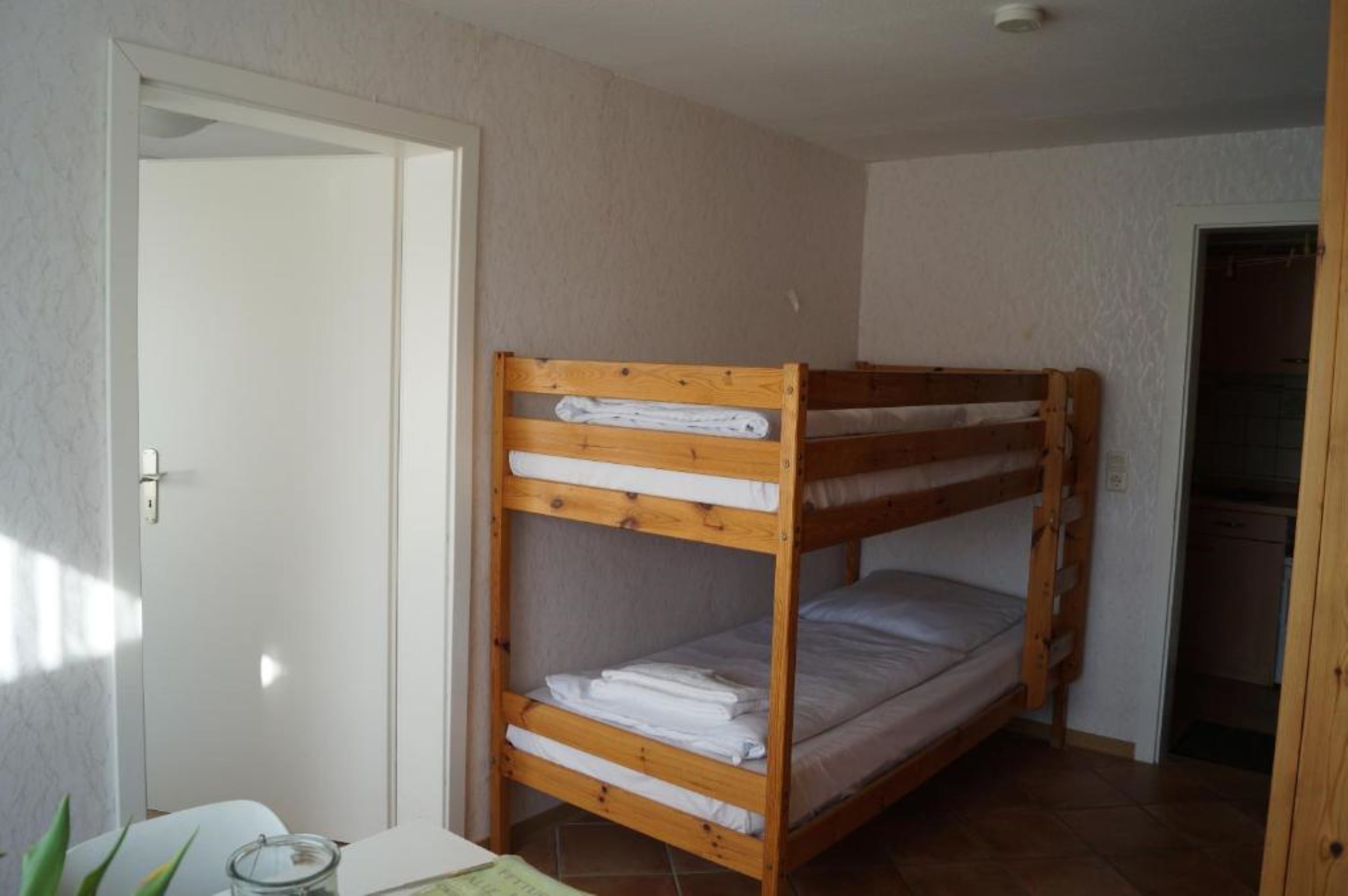 Een stapelbed in een kamer met een witte deur, een klein tafeltje met een glazen vaas en een keukenruimte zichtbaar door een geopende deur.