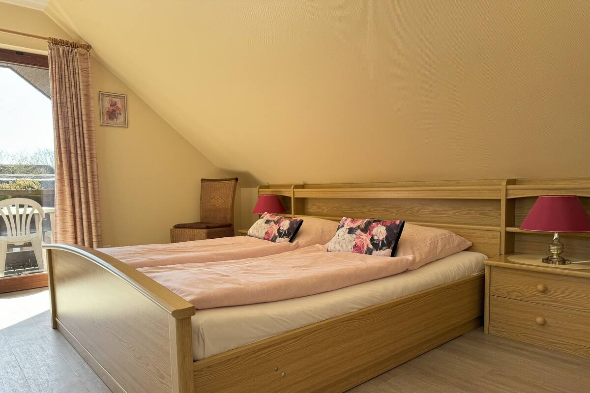 Un lit simple avec une tête de lit en bois, une table de chevet avec une lampe et une chaise près d’une fenêtre avec des rideaux.
