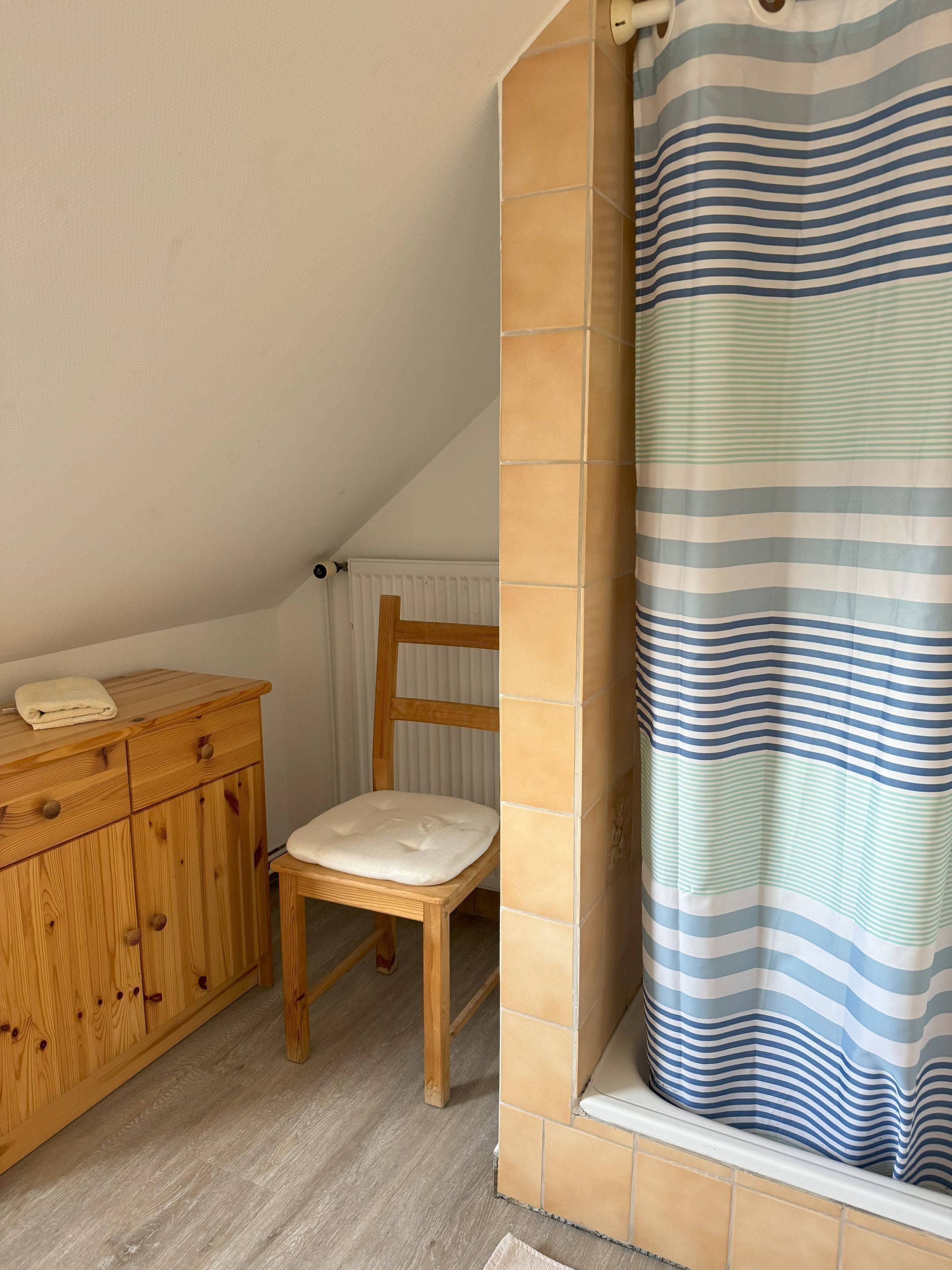 Een kleine, knusse kamer met een houten kast, een comfortabele stoel en een kamerbadgordijn met strepen.