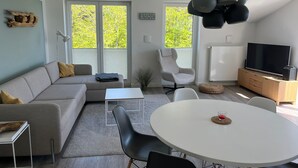 Una sala de estar moderna con sofá, mesa de comedor, sillas, televisor y ventanas amplias.
