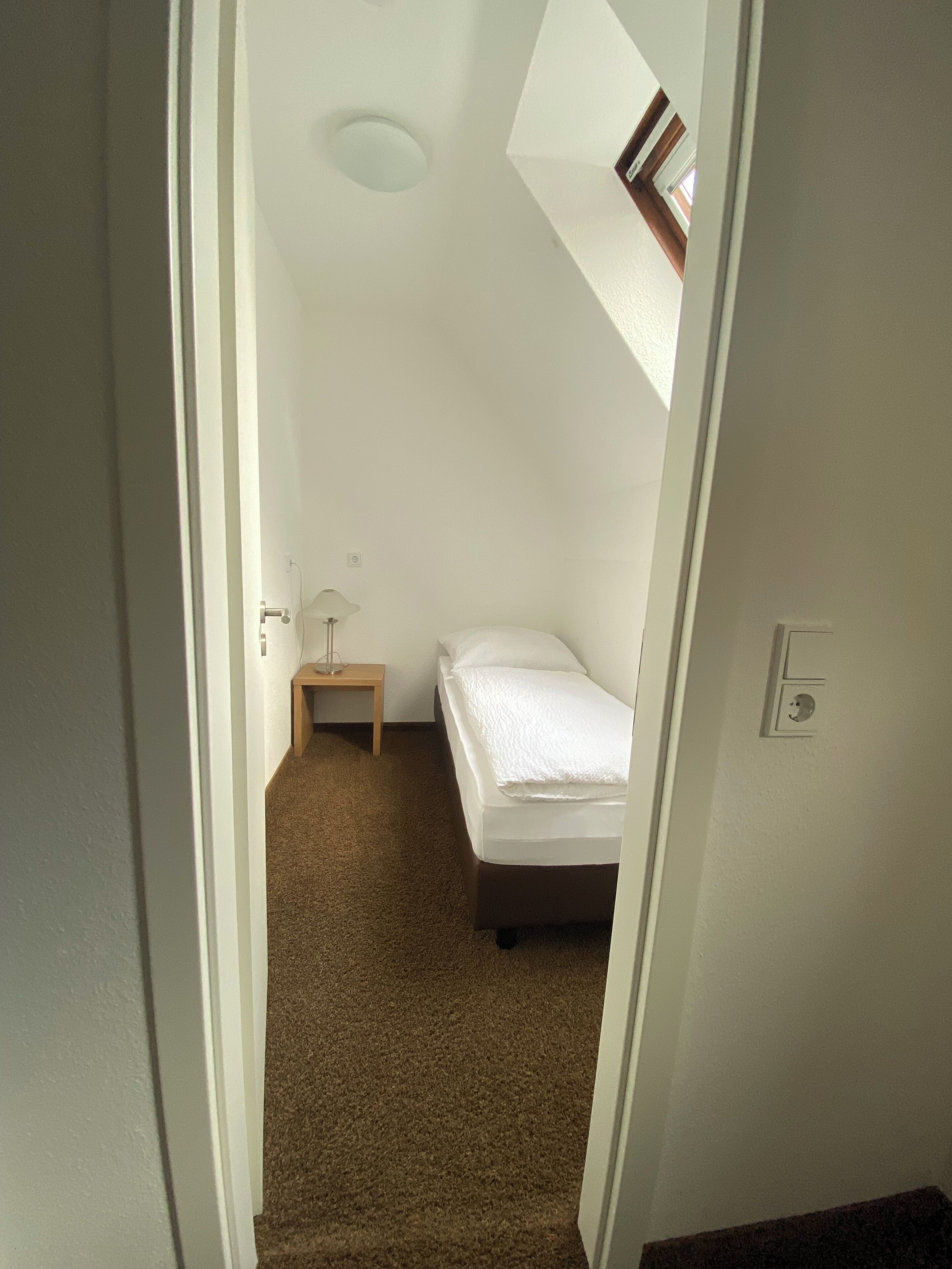 A small, narrow room with a sloped ceiling, a single bed, a bedside table, and a lamp.