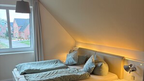 Une chambre à coucher avec un lit, des tables de chevet, une lampe, une fenêtre et une vue sur les maisons à l’extérieur.