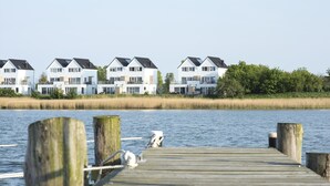 A wooden pier leading to a row of modern white houses with dark roofs, overlooking a calm body of water.