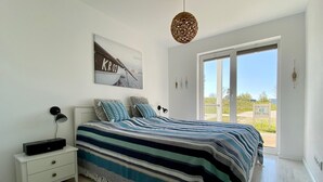 A bedroom with a large bed, a nightstand, a wall-mounted lamp, a hanging light fixture, and a window with a view of the outside.