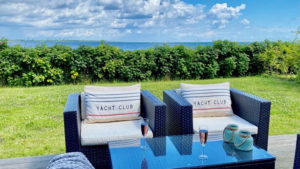 A wicker outdoor seating area with two cushions labeled "YACHT CLUB," a glass table with two champagne flutes, and a view of the sea.