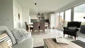 A modern living room with a sofa, coffee table, and a black armchair. There is a dining area with a table and chairs, and a kitchen in the background.