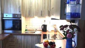 Une cuisine moderne avec des meubles en bois, un four en acier inoxydable, un évier et un plan de travail agrémenté d’une planche à découper, de légumes et d’un vase de fleurs.
