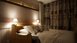 Et hotelværelse med sengegavl i træ, to sengeborde med lamper, en seng med hvide sengetøj og puder med mønster, samt et gardin med detaljeret design.