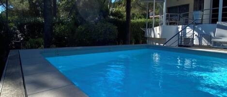 A swimming pool with a wooden deck, surrounded by palm trees and a covered area with lounge chairs.