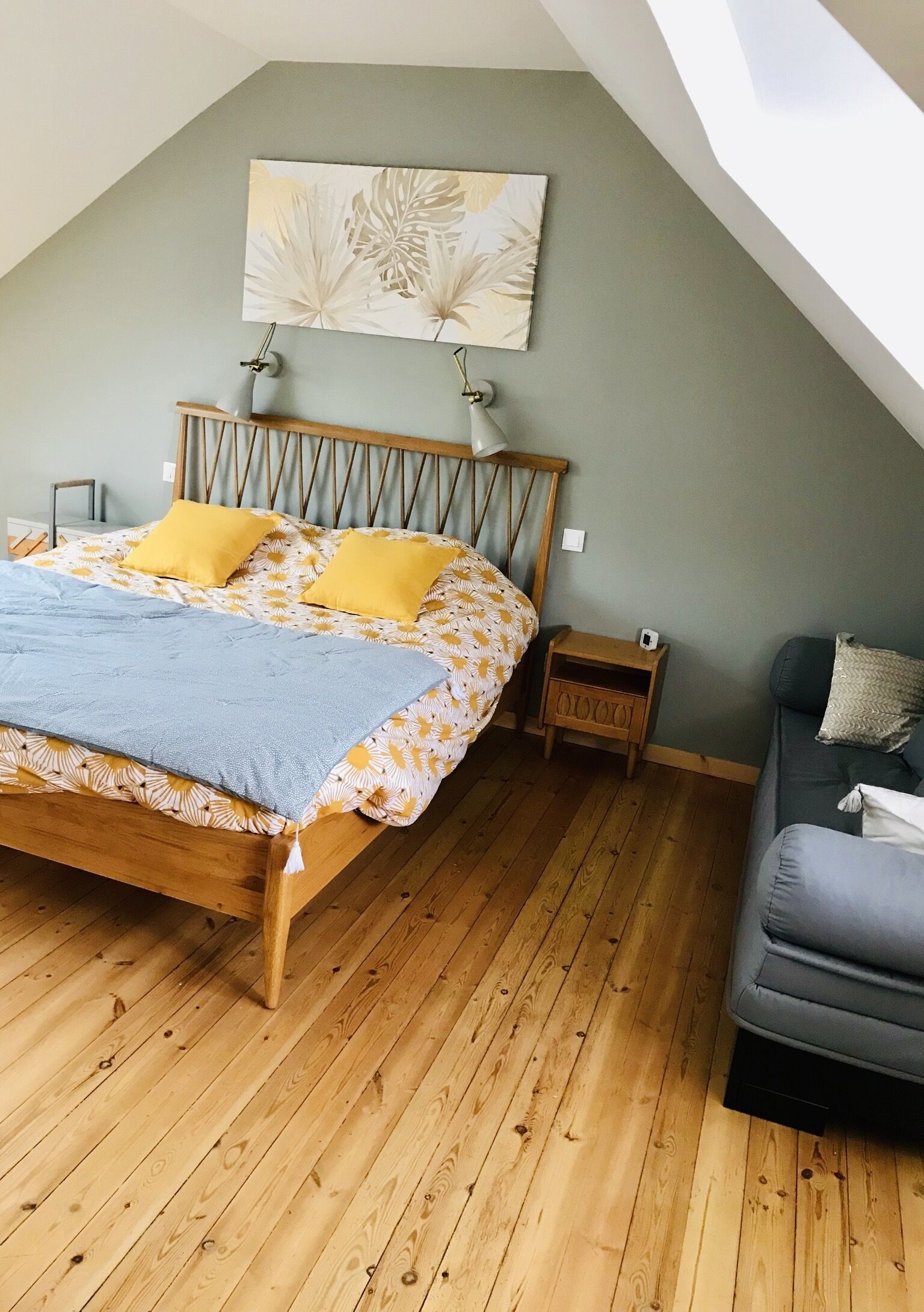 Un dormitorio con una cama de madera, un mesita de noche, un sofá gris y un techo con una obra de arte con diseño de hojas.
