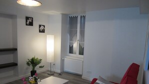 Una sala de estar moderna con un sofá blanco, un sofá rojo, una mesa de centro blanca y una planta. Hay dos marcos de cuadros en la pared y una ventana con cortinas.