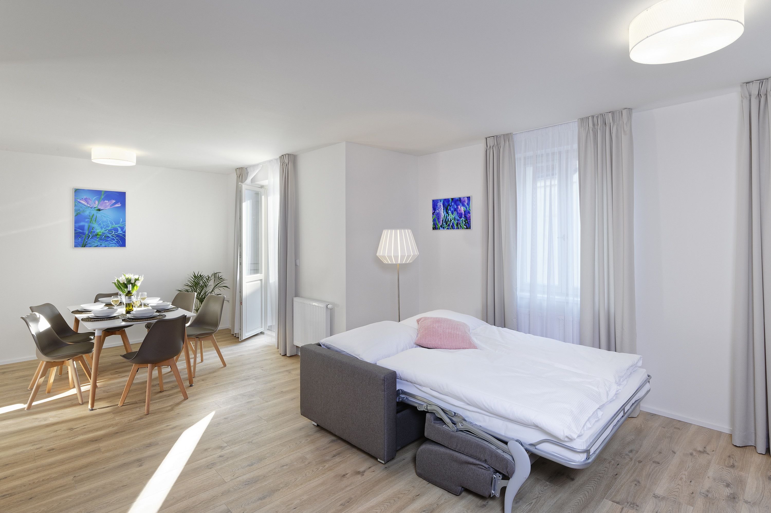 A modern hotel room with a bed, bedside table, lamp, and a dining area with chairs and a table.
