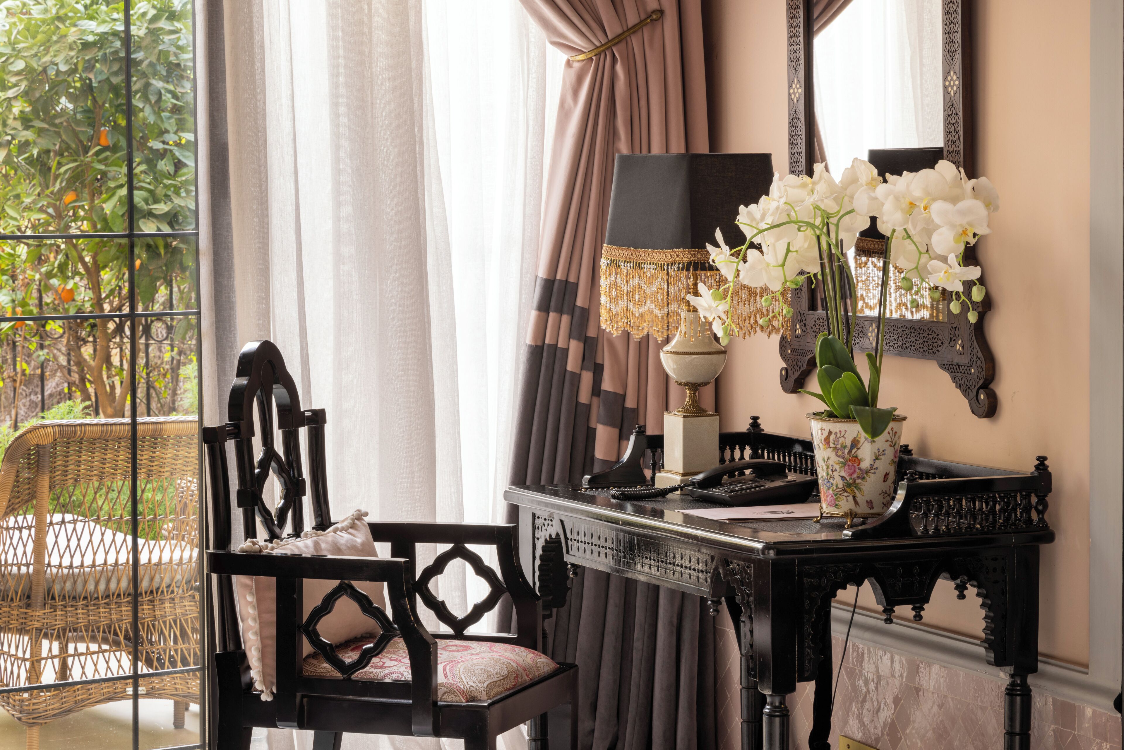 A room with a wicker chair, a desk with a lamp, a vase with flowers, and a mirror on the wall.