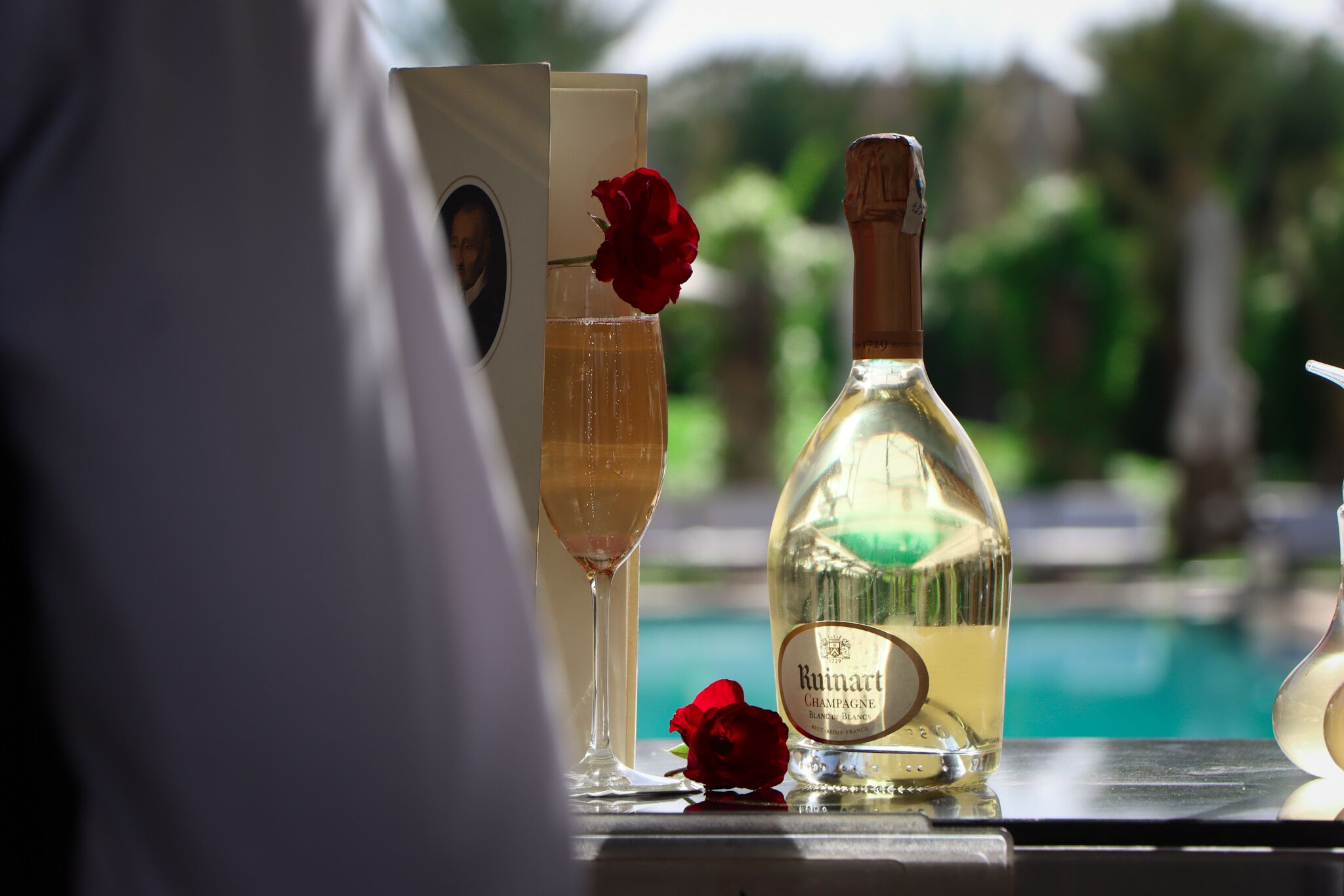 A bottle of Ruivart Champagne Blanc and a glass of champagne with a rose on a table by a pool.