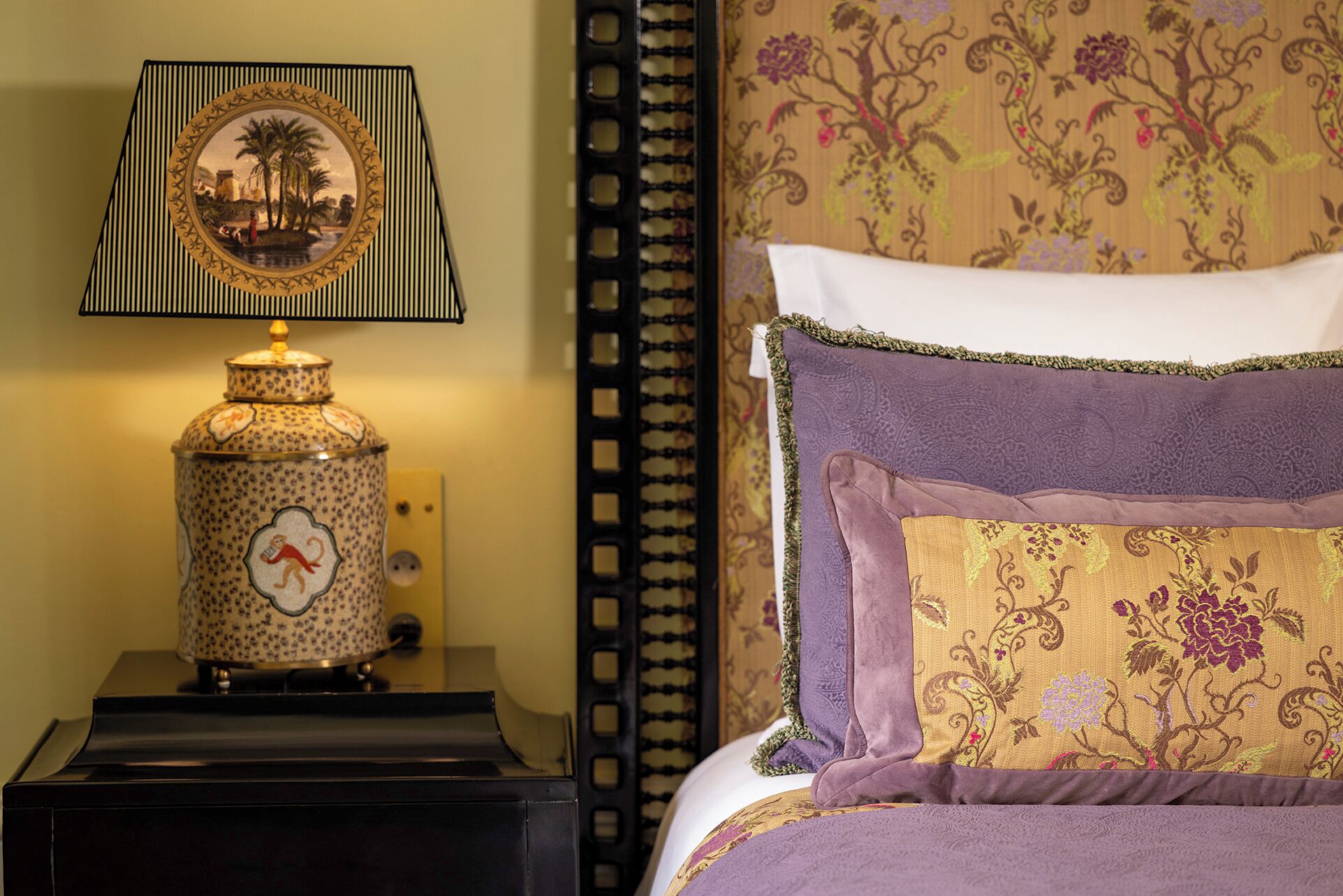 Una cama con edredón y almohadas estampadas con motivos florales, una lámpara decorativa con diseño de palmera y una pared con estampado.