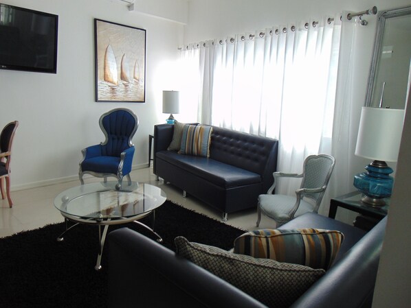 Uma sala de estar com um sofá, poltronas, uma mesa de centro de vidro e um quadro na parede.