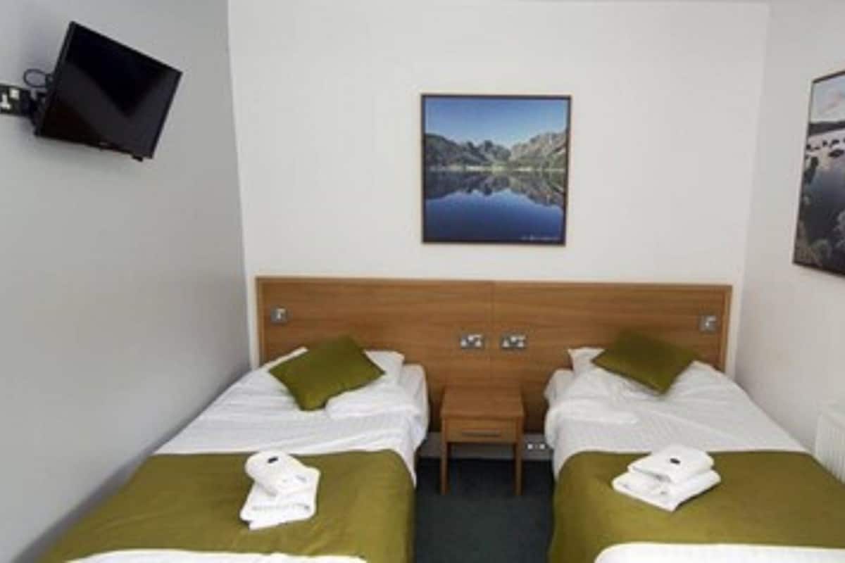 A hotel room with two single beds, a wooden headboard, a flat-screen TV mounted on the wall, and two framed pictures depicting landscapes.